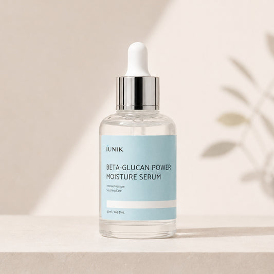 A clear glass dropper bottle of IUNIK Beta-Glucan Power Moisture Serum with a light blue label and a silver dropper cap, set on a light beige surface with soft shadows and leafy shadows in the background.